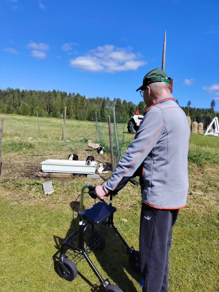 Henkilö seisoo rollaattorin kanssa aurinkoisessa maalaismaisemassa.