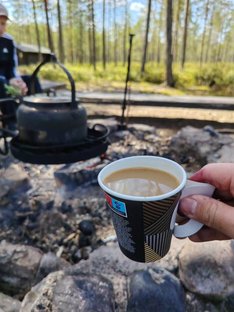 Käsi pitää pahvista kuppia jossa on kahvia. Taustalla kahvipannu ja metsää.