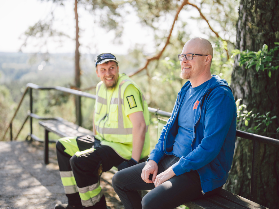 Mies työvaatteissa, joissa heijastinraitoja ja huomiovärejä istuu toisen miehen, jolla Onnikodin ohjaajan työvaatteet kanssa penkillä, juttelevat
