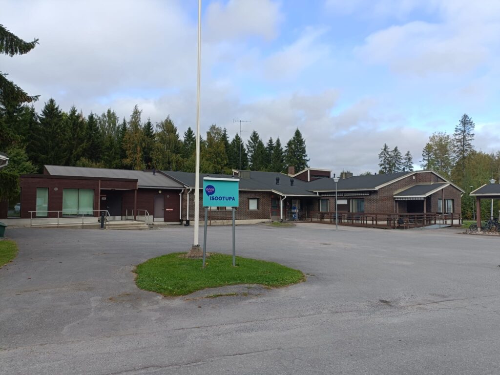 Onnikoti Isoo-tupa-kyltti onnikodin pihalla, iso ruskea rakennus, lipputanko