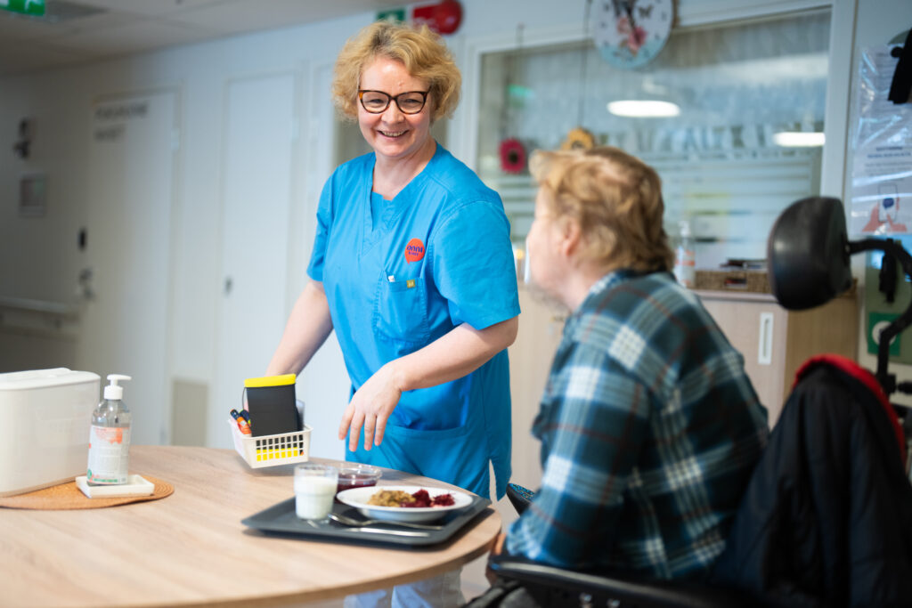 Onnikodin työntekijä ruokapöydän ääressä hymyillen asukkaalle joka aloittamassa ateriaa. Ruoka-annos ja ruokapöytä.