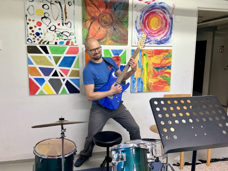 Onnikotien ohjaaja Jussi soittamassa kitaraa bändihuoneessa, värikkäitä tauluja