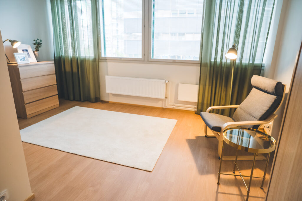 Asukashuone, jossa valkoinen matto, vihreät verhot, puinen lipasto ja harmaa nojatuoli.