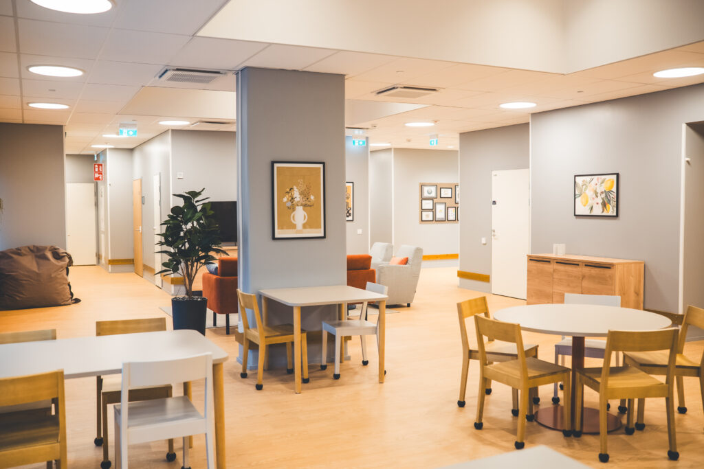 Yhteinen oleskelutila, ruokailuryhmiä, tauluja seinillä, huonekasvi.