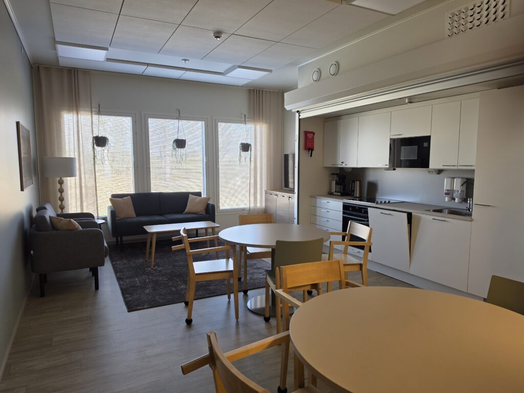 Yhteinen tila, jossa on keittiö, pöytiä, sohvia ja suuret ikkunat.