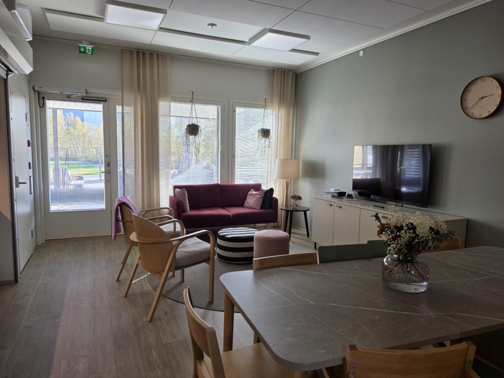 Yhteinen oleskelutila, jossa on sohva, ruokapöytä, televisio ja suuret ikkunat