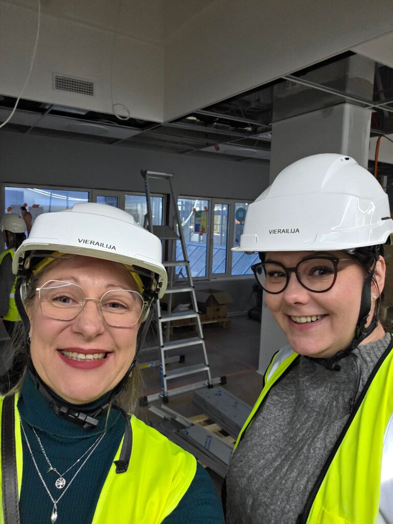 Yksikönjohtaja ja palvelujohtaja hymyilevät työmaakierroksella kypärät päässä, taustalla rakentuva Onnikoti